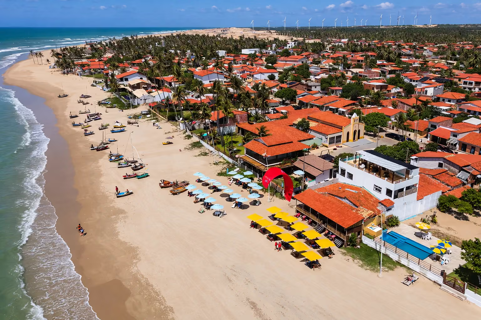 Vista aérea da vila de Guajiru, com barcos de pesca na areia, guarda-sóis e um kitesurista, turbinas de vento ao fundo