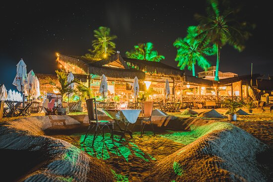 Mesas cavadas na areia em um recanto da praia de Flecheiras iluminado à noite, com coqueiros sob luz verde