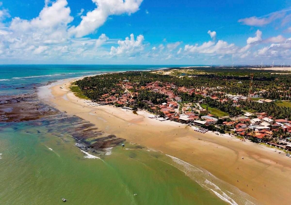 Vista aérea da vila de Flecheiras, com a praia, os recifes, os coqueirais e turbinas de vento ao fundo