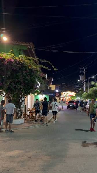 Rua da vila de Flecheiras à noite, com árvores floradas, lojas e movimento
